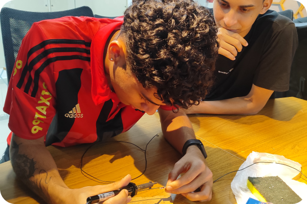 Dois jovens estão trabalhando juntos; um está conectando fios a um sensor enquanto o outro observa atentamente.