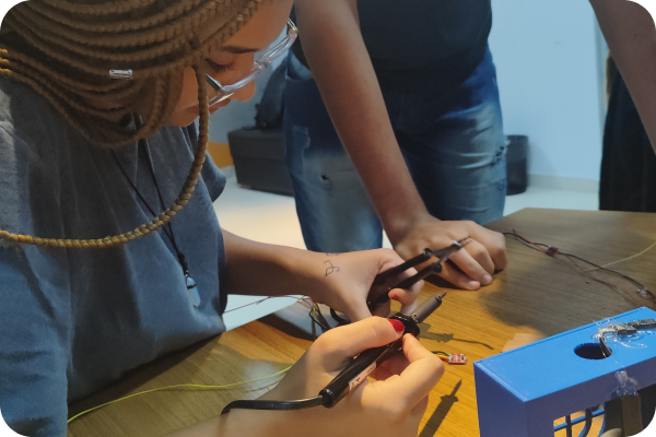 Uma jovem realiza uma soldagem, enquanto uma figura em pé ao seu lado permanece fora do foco, sem mostrar o rosto.
