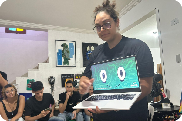 Uma mulher em pé segura um notebook, apresentando a imagem do rostinho de Chico sorrindo para um grupo de pessoas sentadas ao seu redor.